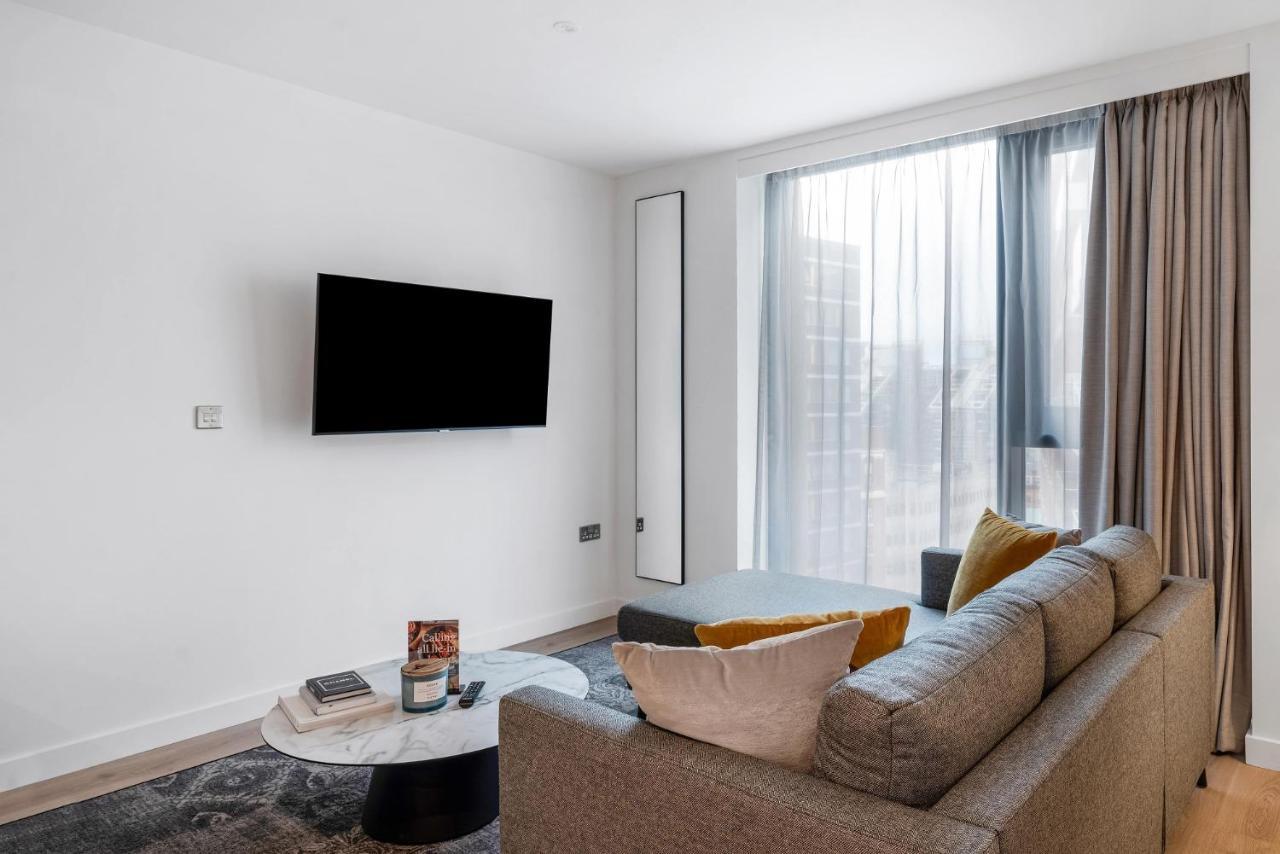 Citysuites 2 Aparthotel Salford  Exterior foto The image shows a modern living room with a minimalist design. There is a large flat-screen TV mounted on the white wall. A comfortable gray sofa with textured cushions occupies the center of the room, complemented by a round coffee table in front of