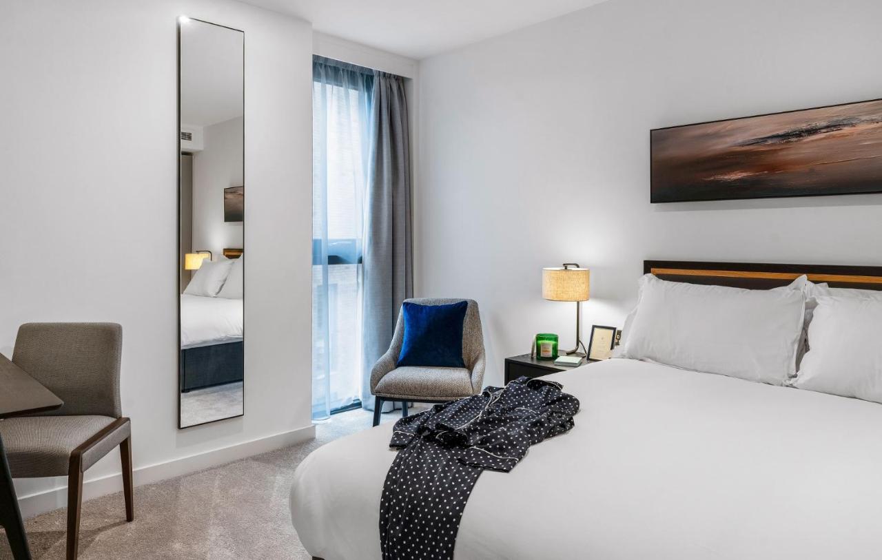 Citysuites 2 Aparthotel Salford  Exterior foto The photo shows a modern hotel room. It features a neatly made bed with white linens and a dark-colored throw draped over it. There is a small bedside table with a lamp next to the bed. A comfortable chair in a light color is positioned near the wind