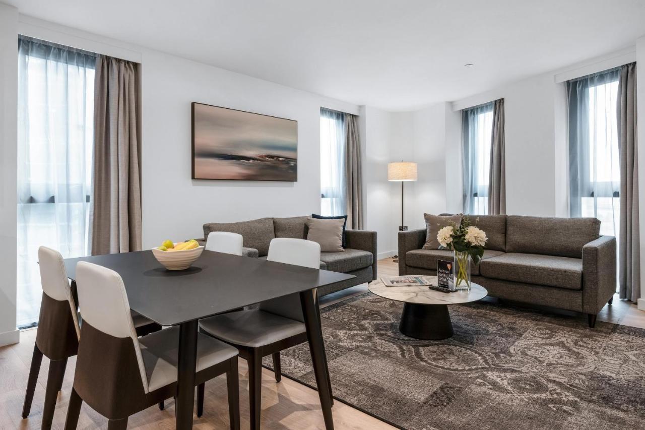 Citysuites 2 Aparthotel Salford  Exterior foto The photo depicts a modern and stylish interior of a living space. On the left, there is a dining table with four chairs, featuring a bowl of fruits on the table. In the background, there's a cozy seating area with a gray sofa and a smaller round cof