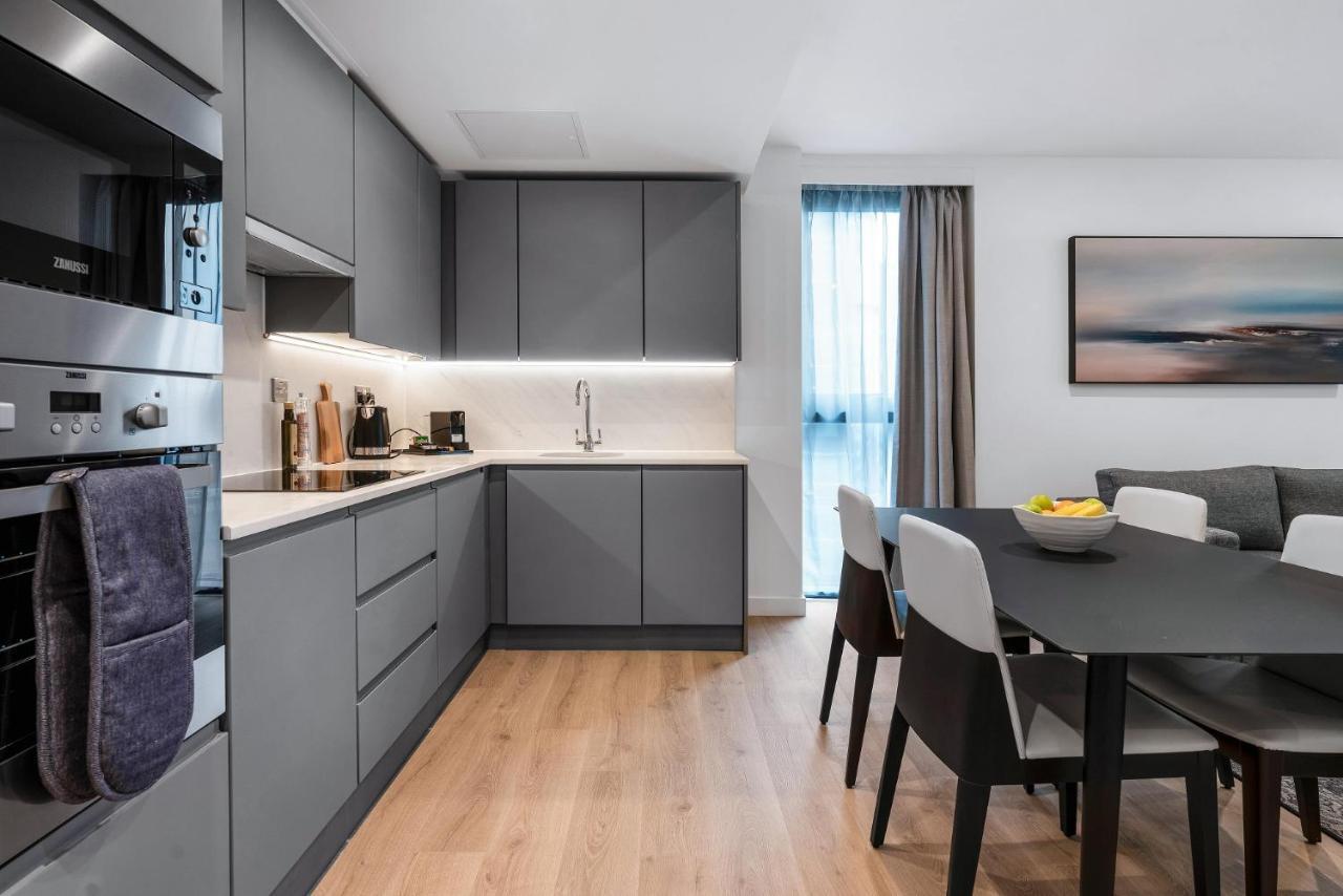 Citysuites 2 Aparthotel Salford  Exterior foto The photo shows a modern kitchen and dining area. The kitchen features sleek gray cabinetry, a white countertop, and built-in appliances such as an oven. There is a sink and some kitchen utensils displayed. The adjacent dining area contains a black t