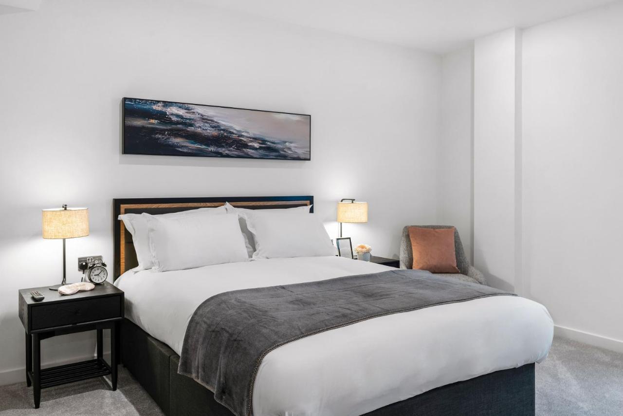 Citysuites 2 Aparthotel Salford  Exterior foto The photo shows a modern bedroom featuring a large bed with crisp white linens and a grey throw blanket. On either side of the bed, there are bedside tables with lamps. A piece of abstract art hangs above the headboard, and there is a chair in a corn