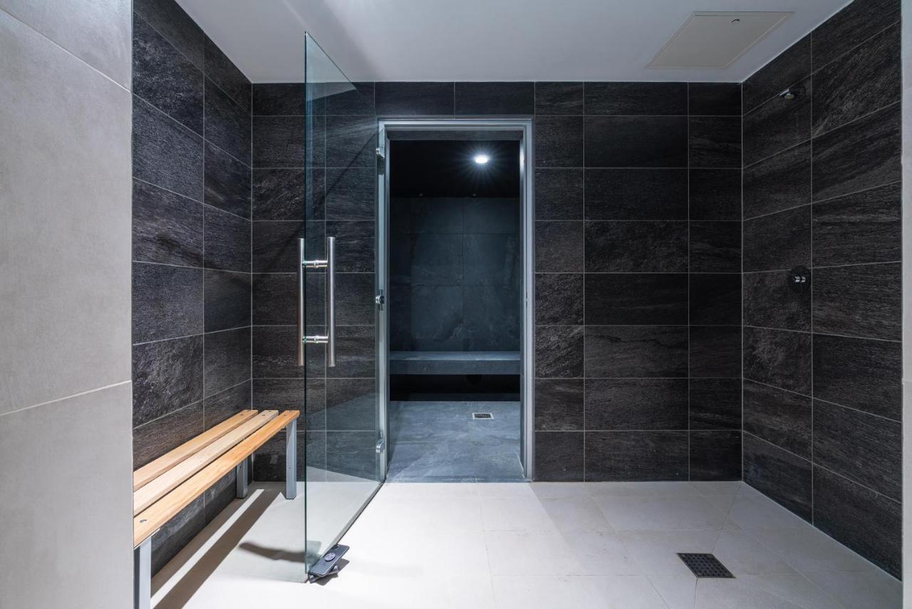 Citysuites 2 Aparthotel Salford  Exterior foto The photo shows a modern, minimalist shower area. The space is predominantly tiled with dark gray tiles on the walls, giving it a sleek and sophisticated look. There is a glass door that leads to another room, and a wooden bench is positioned against